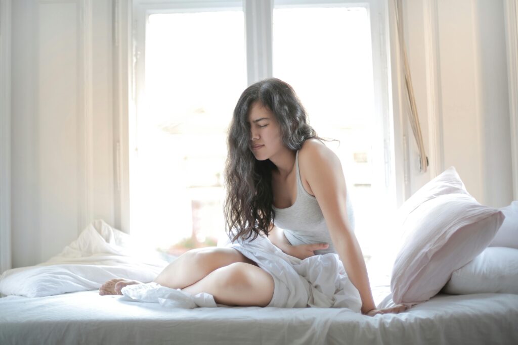 pexels-photo-3807733-3807733 Woman sitting on bed holding her stomach, showing signs of discomfort or pain indoors.